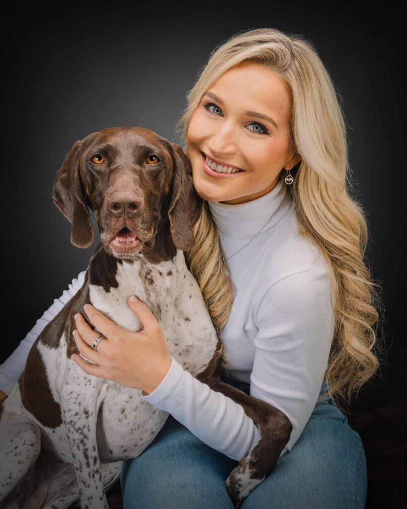 Woman with blonde hair and white shirt with gsp dog smiling in studio with black backdrop